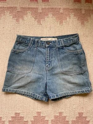 Blå denimshorts från Nevada - Klassiska blå denimshorts från Nevada med medel hög midja och raka ben. Shortsen har cargo stil med stora framfickor, bakfickor med knappdetaljer och bälteshällor. Perfekta till sommaren och gjorda i slitstark bomull.