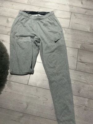 Gråa mjukisbyxor från Nike - Säljer ett par gråa mjukisbyxor från Nike med resår i midjan och smal passform. Byxorna har sidofickor och en svart Nike-logga på vänster ben. Tillverkade i mjukt och bekvämt material, perfekta för chill dagar.