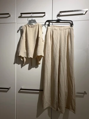 Beige set med topp och vida byxor - Säljer ett snyggt tvådelat set i beige med en ärmlös topp som knyts framtill och matchande vida byxor med resår i midjan. Materialet är mjukt och har en lätt skrynklig struktur som ger en avslappnad look. 