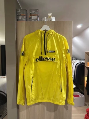 Gul vindjacka från Ellesse - Snygg gul vindjacka från Ellesse med stor logga på bröstet och mindre logga på ärmen. Jackan har huva med dragsko, halv dragkedja framtill och en stor magficka med lock. Tillverkad i lätt och glansigt material, perfekt för blåsiga dagar.