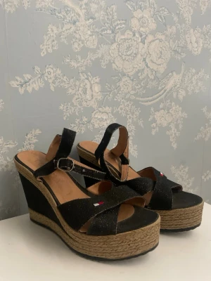 TOMMY HILFIGER Svarta kilklackade sandaler - Snygga svarta TOMMY HILFIGER sandaler med hög kilklack och flätad sula i rep. Remmarna har ett litet broderat flaggmärke och justerbart spänne vid ankeln. Ovandelen är i textil och sulan har en trendig espadrille-look. Perfekta för dig som vill ha extra höjd och stil. 
