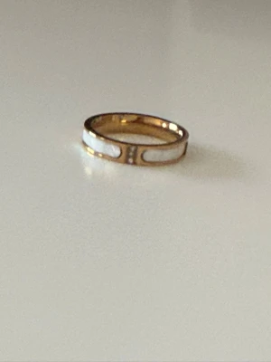 Guldfärgad ring i Rostfritt stål med vit emalj - Snygg guldfärgad ring i rostfritt stål med vita emaljdetaljer och små glittrande stenar framtill. Ringen har en modern, stilren design med öppna partier och passar perfekt för dig som gillar trendiga accessoarer.storlek 16,5mm
