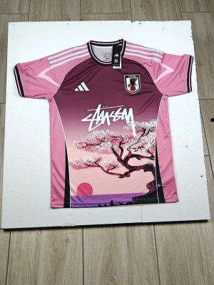 Rosa Japan fotbollströja Stussy x Adidas - Unik rosa fotbollströja med Japan-emblem och Stussy-logga framtill. Tröjan har ett snyggt körsbärsblomsmotiv och vita detaljer på axlarna. Tillverkad i lätt och ventilerande polyester, perfekt för träning eller match.