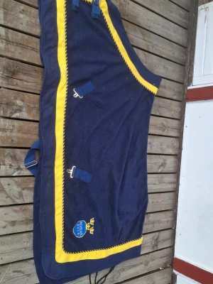 Blå hästtäcke med gula detaljer - Mörkblå hästtäcke med gula kanter och broderade detaljer,. Täcket har metallspännen och remmar för enkel fastsättning. Perfekt för att hålla hästen varm och snygg i stallet.
