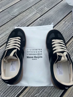 Maison Margiela svarta sneakers - Maison Margiela sneakers i svart läder och mocka med vita snören och beige innersula. Klassisk låg modell med gummisula i brunt och diskreta detaljer. Perfekta för dig som gillar stilrena och exklusiva sneakers.