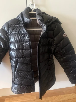 Svart dunjacka från Colmar med huva - Snygg svart dunjacka från Colmar med quiltad design och huva. Jackan har dragkedja framtill och en logotyp på ärmen