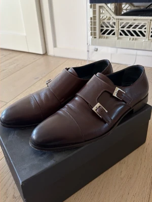 Bruna dubble monkstrap loafers (Cacharel) - Snygga bruna loafers med dubbla monkstrap-spännen i silverfärg. Skorna har en elegant, spetsig tå och är tillverkade i slätt läder. Använda 1 gång. 