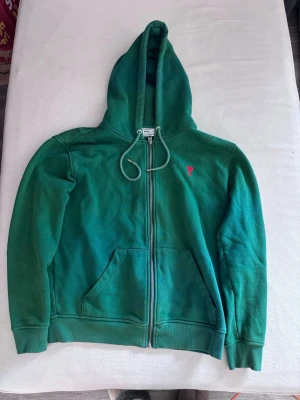 Ami Paris hoodie - Snygg grön hoodie från Ami Alexandre Mattiussi med dragkedja framtill, huva med dragsko och kängurufickor. Liten röd broderad detalj på bröstet. Tillverkad i mjuk bomull för skön känsla. Perfekt för en chill och avslappnad stil.