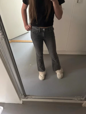 Grå bootcut jeans med detaljer - Säljer ett par grå bootcut jeans med snygga detaljer på bakfickorna. Jeansen har låg midja, klassisk femficksmodell och är tillverkade i stretchigt denimtyg. Perfekta för dig som gillar en avslappnad men trendig look.