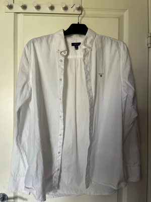 Vit skjorta från Gant med knappar - Klassisk vit skjorta från Gant med knappstängning och broderad logga på bröstet. Skjortan har lång ärm, normal passform och är tillverkad i bomull. Perfekt till många olika stilar och enkel att matcha.