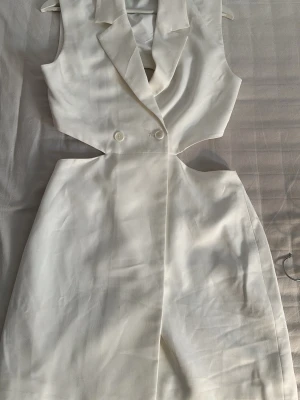 Vit omlottklänning med cut-outs - Säljer en vit kortklänning med omlottdesign och cut-outs i midjan. Klänningen har krage, dubbla knappar framtill och är ärmlös. Perfekt för dig som vill ha en modern och stilren look.