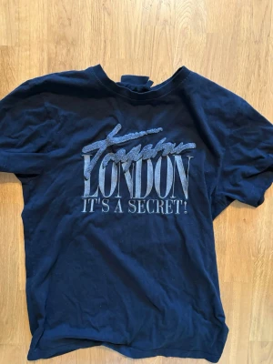 Trapstar t shirt - Snygg svart T-shirt med stort tryck på bröstet där det står 'London It's a Secret!'. Klassisk rund hals och korta ärmar. Perfekt för dig som gillar streetstyle och vill ha en cool statement-tee i garderoben.
