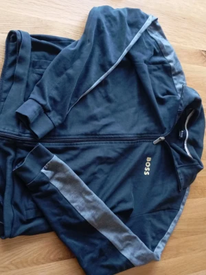 Svart boss tröja - Snygg svart elegant tröja från BOSS med dragkedja framtill och gråa detaljer längs ärmarna. Klassisk passform med ribbade muddar och diskret BOSS-logga på bröstet. Perfekt för en avslappnad och stilren look.