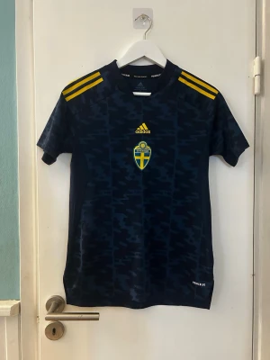 Svensk fotbollströja Adidas blå - Mörkblå fotbollströja från Adidas med guldfärgade detaljer och Sveriges landslagsemblem på bröstet. Tröjan har korta ärmar, rund hals och gula ränder på axlarna. Tillverkad i lätt och ventilerande material, perfekt för träning eller match. Använd en gång