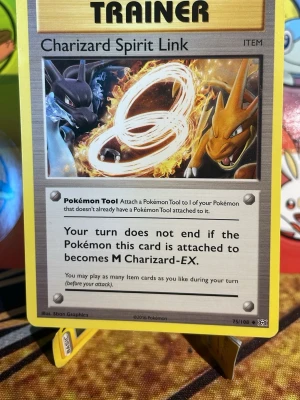 Pokémon Charizard Spirit Link kort - Charizard Spirit Link är ett Pokémon TCG-tränarkort med motiv av Charizard och Mega Charizard. Kortet har en gul ram, färgstark illustration och text på engelska. Perfekt för samlare eller dig som vill boosta din lek med ett unikt itemkort.