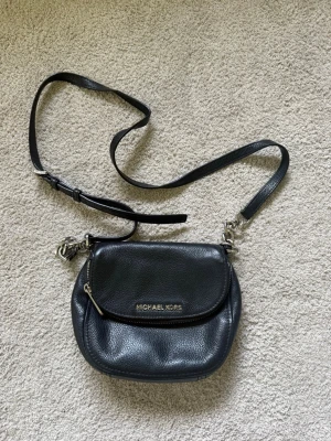 Svart axelväska Michael Kors - Snygg svart axelväska från Michael Kors i mjukt läder med justerbar axelrem och silverfärgade metalldetaljer. Väskan har ett lock med dragkedja och logga framtill. Även ett extra fack i väskan med dragkedja. Den lilla remmen som håller fast kedjan har lossnat på ena sidan, men den går enkelt att sy fast igen. I övrigt fint skick.