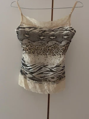  linnetopp med spets - Snygg linnetopp med tunna axelband och mixade djurmönster i beige och svart. Toppen har detaljer i spets upptill och nedtill samt inslag av leopard- och zebramönster. Perfekt för dig som gillar unika prints och en feminin touch.