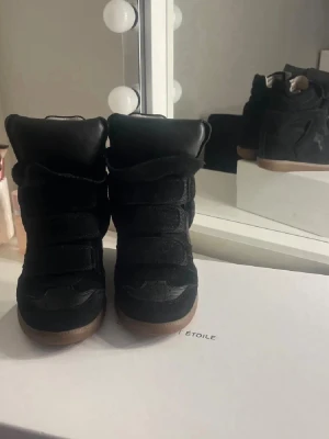 Svarta sneakers med kilklack - Snygga svarta sneakers med kilklack och kardborreband. Skorna har en blandning av mocka och läderdetaljer samt en brun gummisula. Perfekta för dig som vill ha lite extra höjd och en cool streetstil.
