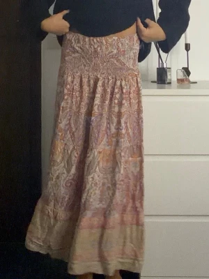 Långkjol med paisleymönster i pastell - Bohemisk långkjol med paisleyinspirerat mönster i ljusa toner av rosa, beige, orange och lila. Kjolen har smock i midjan för skön passform och är tillverkad i ett lätt, flödande material som passar perfekt till vår och sommar.