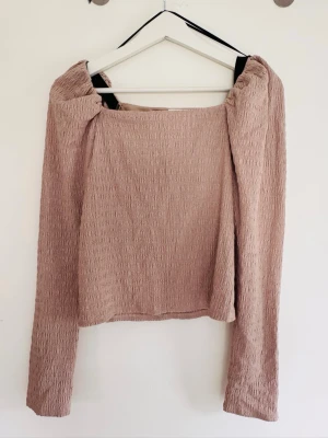 Beige långärmad topp från H&M - Trendig beige långärmad topp från H&M med smockad struktur och fyrkantig ringning. Toppen har en croppad passform och är tillverkad i ett mjukt, stretchigt material som känns bekvämt mot huden. Perfekt att styla med höga jeans eller kjol.