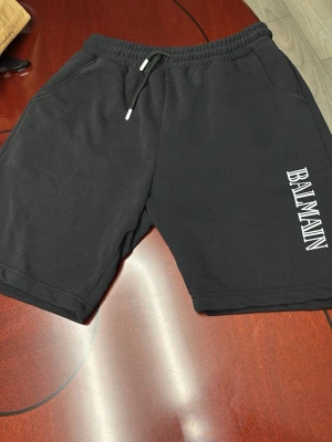 Balmain Shorts - Säljer ett par svarta shorts från Balmain med vit logga på ena benet. Shortsen har elastisk midja med snörning och sidofickor. Perfekta för en avslappnad och trendig stil.