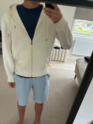 Creme hoodie med dragkedja från Polo Ralph Lauren - Snygg Creme hoodie från Polo Ralph Lauren med dragkedja framtill och klassisk huva med dragsko. Broderad logga i beige på bröstet. Tillverkad i mjukt bomullsmaterial och har ribbade muddar vid ärmslut och nederkant. Perfekt för en clean och stilren look.