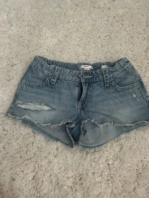 Blå jeansshorts med paljettdetaljer - Säljer ett par blå jeansshorts med slitna kanter och en snygg reva framtill. Baktill har shortsen coola fickor med silverfärgade paljetter i ett mönster. Perfekta för sommaren och riktigt trendiga till alla outfits.