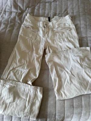 Beiga jeans  med fickor - Snygga beiga byxor med flera stora fickor framtill och detaljerade sömmar. Byxorna har raka ben och klassisk stil  med lock på fickorna. Tillverkade i ett slitstarkt bomullstyg som passar perfekt till en avslappnad look.