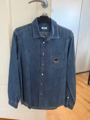 Blå jeansskjorta från Kenzo - Säljer en klassisk blå jeansskjorta från Kenzo med lång ärm och knappar framtill. Skjortan har en bröstficka med Kenzo-tigern broderad på. Perfekt till ett avslappnat men snyggt vardagslook. Använd en handfull gånger. Storlek S