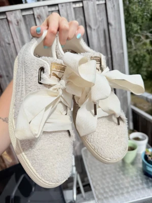 Vita sneakers med fina band från Puma - Unika vita sneakers från Puma med fluffig teddytextur och stora, breda rosett-snören. Skorna har gulddetaljer på plösen och klassisk låg modell. Perfekta för dig som vill sticka ut med en söt och trendig look. Använda en gång! Toppen-skick. 