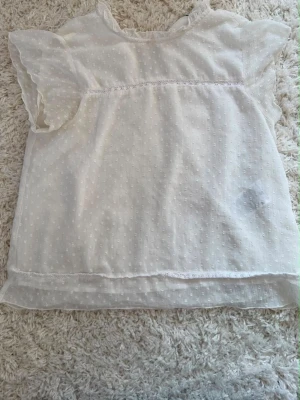 Vit blus med volangärm och prickigt mönster - Söt vit blus med diskret prickigt mönster och volangdetaljer vid ärmarna. Blusen är tillverkad i ett tunt, lätt material och har en liten öppning bak i nacken. Perfekt för en fräsch och luftig look.