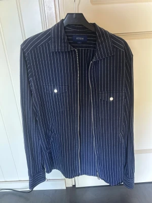 Mörkblå randig overshirt från Eton - Snygg mörkblå overshirt från Eton med vita vertikala ränder. Modellen har två bröstfickor med knappar, dragkedja framtill och klassisk krage. Tillverkad i ett mjukt tyg som känns lyxigt och stilrent. Perfekt för lager på lager och en clean look.