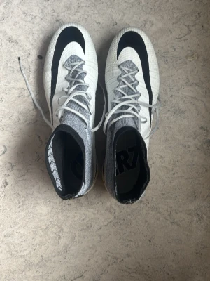 Vita CR7 fotbollsskor med högt skaft - Säljer ett par vita fotbollsskor med högt skaft och CR7-detaljer. Skorna har svart Nike-logga, gråmelerad strumpa och gula detaljer vid sulan. Ovandelen är i syntetmaterial med snörning och dobbar för spel på gräs.