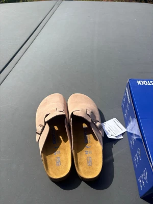  Birkenstock Boston Mud - Säljer ett par snygga beige tofflor från Birkenstock i mjuk mocka. Modellen har en klassisk slip-in design med justerbart spänne och ergonomisk fotbädd för extra komfort. Perfekta för chill dagar hemma eller när du vill ha en avslappnad stil.
