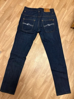 Mörkblå Nudie Jeans - Säljer ett par mörkblå jeans från Nudie Jeans med klassisk femficksmodell och kontrastsömmar. Jeansen har raka ben och tydliga broderade detaljer på bakfickorna. Tillverkade i slitstark denim, perfekta för en clean och stilren look.