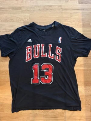Nba Chicago Bulls t-shirt Noah 13 - Svart t-shirt från Adidas med Chicago Bulls-tryck och nummer 13 på både fram- och baksida. På ryggen står det NOAH och Bulls-loggan syns ovanför. T-shirten är kortärmad och har röda och vita detaljer i trycket. Perfekt för basketfans! Obs en litet hål mellan 1 och 3