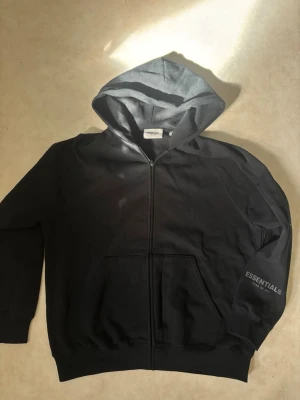 Svart Essentials hoodie med dragkedja oversized - Svart hoodie från Essentials med dragkedja framtill, huva och fickor. Diskret Essentials-logga på ärmen och etikett på huvan. Tillverkad i mjukt material som passar perfekt till en avslappnad stil. Ej använd storlek M men oversized så passar L meråt med en baggy look