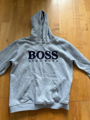 Grå hoodie från Hugo Boss - Snygg grå hoodie från Hugo Boss med stor svart BOSS-logga på bröstet och klassisk känguruficka framtill. Hoodien har huva med dragsko och ribbade muddar vid ärmar och nederkant. Perfekt för en avslappnad och trendig look.