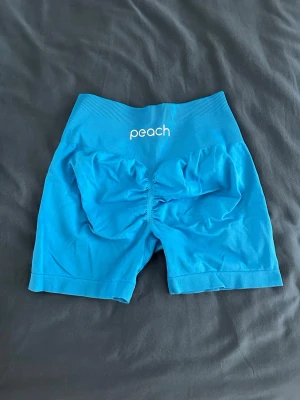 Blå höga seamless shorts Peach - Snygga blå seamless shorts från Peach med hög midja och scrunch-detalj bak. Shortsen har en ribbad linning och är tillverkade i ett stretchigt material som passar perfekt till träning. Färgen är en nyans mellan de två framträdande på bilderna. Klarblå nyans. Storlek S. Sparsamt använda. 