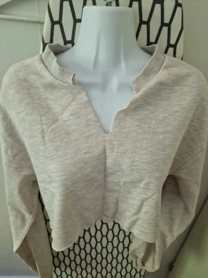 Beige croppad topp med v-ringning - Trendig beige croppad sweatshirt med unik v-ringad hals och ribbad kant. Långärmad modell i mjukt sweatshirtmaterial, perfekt för en avslappnad och cool stil. Passar till både jeans och sweatpants 