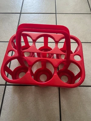 Röd flaskhållare i plast - Praktisk röd flaskhållare i plast med plats för sex flaskor. Perfekt för att bära drycker till picknick, träning eller fest. Har ett rejält handtag i mitten för enkel transport och rundade hål för flaskorna.
