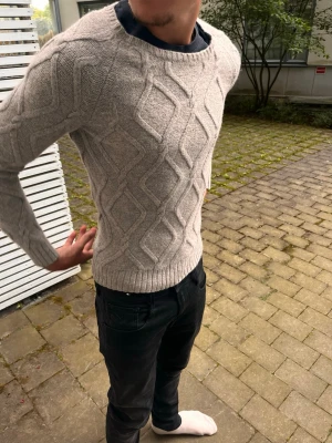 Beige kabelstickad tröja herr - Snygg beige kabelstickad tröja med rund hals och ribbade muddar. Tröjan har ett klassiskt mönster med stora romber och är perfekt för lager-på-lager-stil. Passar dig som gillar en stilren och tidlös look.