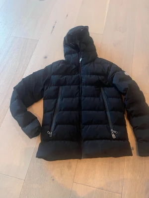 Svart dunjacka med huva från Moncler! - Snygg svart dunjacka med huva och två stora dragkedjeförsedda fickor framtill. Jackan har quiltad design med breda paneler och en hög krage. Perfekt för kalla vinterdagar och ger en stilren look.