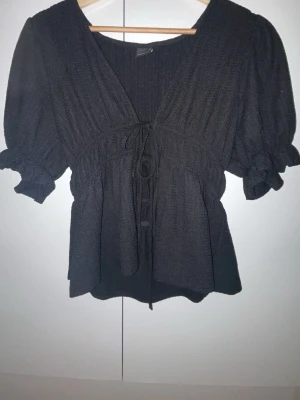 Svart blus med knyt och puffärm - En svart blus med struktur, v-ringning och knytband framtill. Blusen har puffiga korta ärmar med resår och volangdetalj vid ärmslutet. Materialet är lätt och ger ett flowy intryck. Perfekt för dig som gillar en feminin och trendig look.