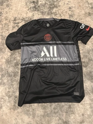 Svart PSG fotbollströja från Nike - Snygg svart PSG matchtröja från Nike med röd Paris Saint-Germain-logga på bröstet och vit 'Accor Live Limitless'-sponsor på magen. Tröjan har gråa detaljer och är tillverkad i Dri-FIT material som andas. Perfekt för träning eller match.