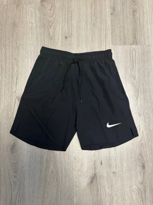 Svarta Nike shorts med snörning - Svarta träningsshorts från Nike med vit logga på benet och snörning i midjan. Shortsen har elastisk midja med Nike-tryck på insidan och är tillverkade i ett lätt och snabbtorkande material. Perfekta för sport och träning.