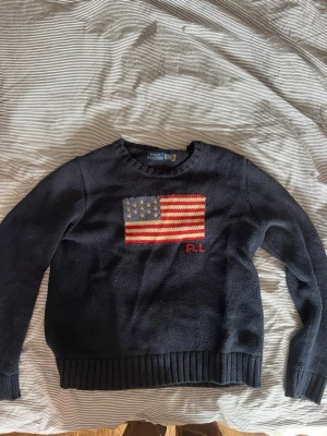 Mörkblå stickad tröja med USA-flagga - Snygg mörkblå stickad tröja med amerikansk flagga på bröstet och RL-broderi i rött. Tröjan har rund hals, långa ärmar och ribbade muddar vid ärmslut och nederkant. Perfekt för dig som gillar klassisk och tidlös stil. Den har krympt lite 