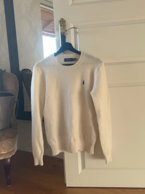 Vit stickad tröja från Polo Ralph Lauren - Klassisk vit stickad tröja från Polo Ralph Lauren med rund halsringning och den ikoniska lilla logotypen broderad på bröstet. Tröjan har ribbade muddar vid ärmslut och nederkant. Perfekt för en clean och stilren look.