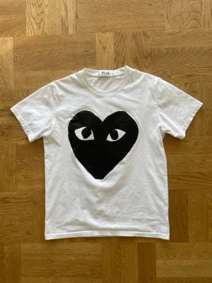 Comme des garçons - Vit t-shirt med ett stort svart hjärta med ögon tryckt på bröstet. Klassisk rund hals och korta ärmar. Perfekt för dig som gillar grafiska prints och vill sticka ut lite extra. Väldigt bra skick! Kan gå ner lite i pris