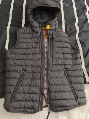 Mörkgrå dunväst från Parajumpers - Snygg mörkgrå dunväst från Parajumpers med huva och dragkedja framtill. Västen har två snedställda fickor med dragkedja och en gul detalj vid halsen. Tillverkad i lättviktsmaterial, perfekt för lager-på-lager och streetwear.